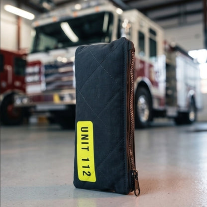 Schwarze, gesteppte Federtasche im Feuerwehr-Design mit gelbem Unit 112 Klettpatch steht aufrecht vor einem roten Löschfahrzeug in einer Fahrzeughalle.