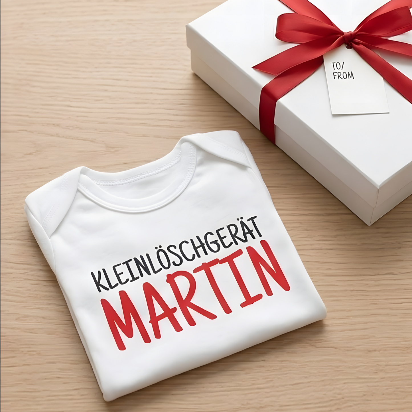Ordentlich gefalteter Feuerwehr-Babybody mit Namensdruck neben einer weißen Geschenkbox mit roter Schleife.