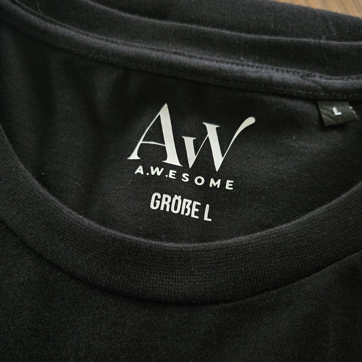Krageninnenseite des schwarzen T-Shirts mit eingedrucktem silbernen AW Awesome Marken-Logo für kratzfreien Tragekomfort.