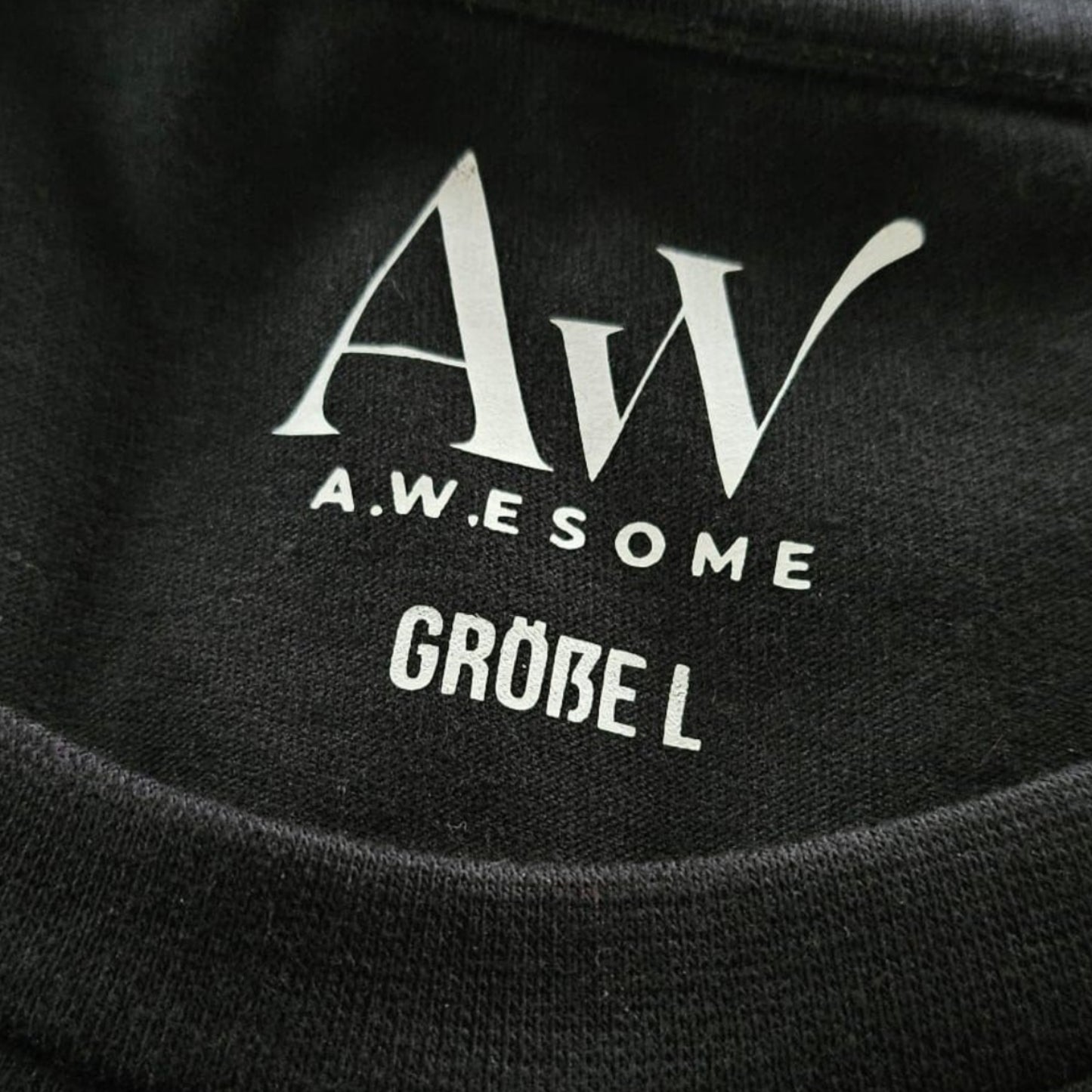 Detail des Nackenbereichs beim schwarzen Pullover mit eingedrucktem weißen AW Awesome Markenlogo und der Größenangabe.