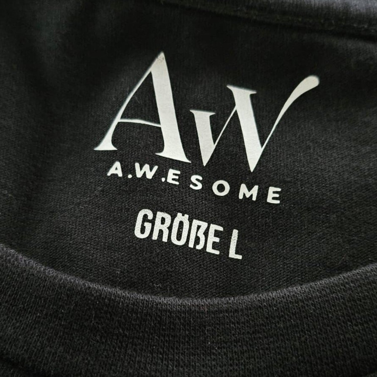 Detail des Nackenbereichs beim schwarzen Pullover mit eingedrucktem weißen AW Awesome Markenlogo und der Größenangabe.