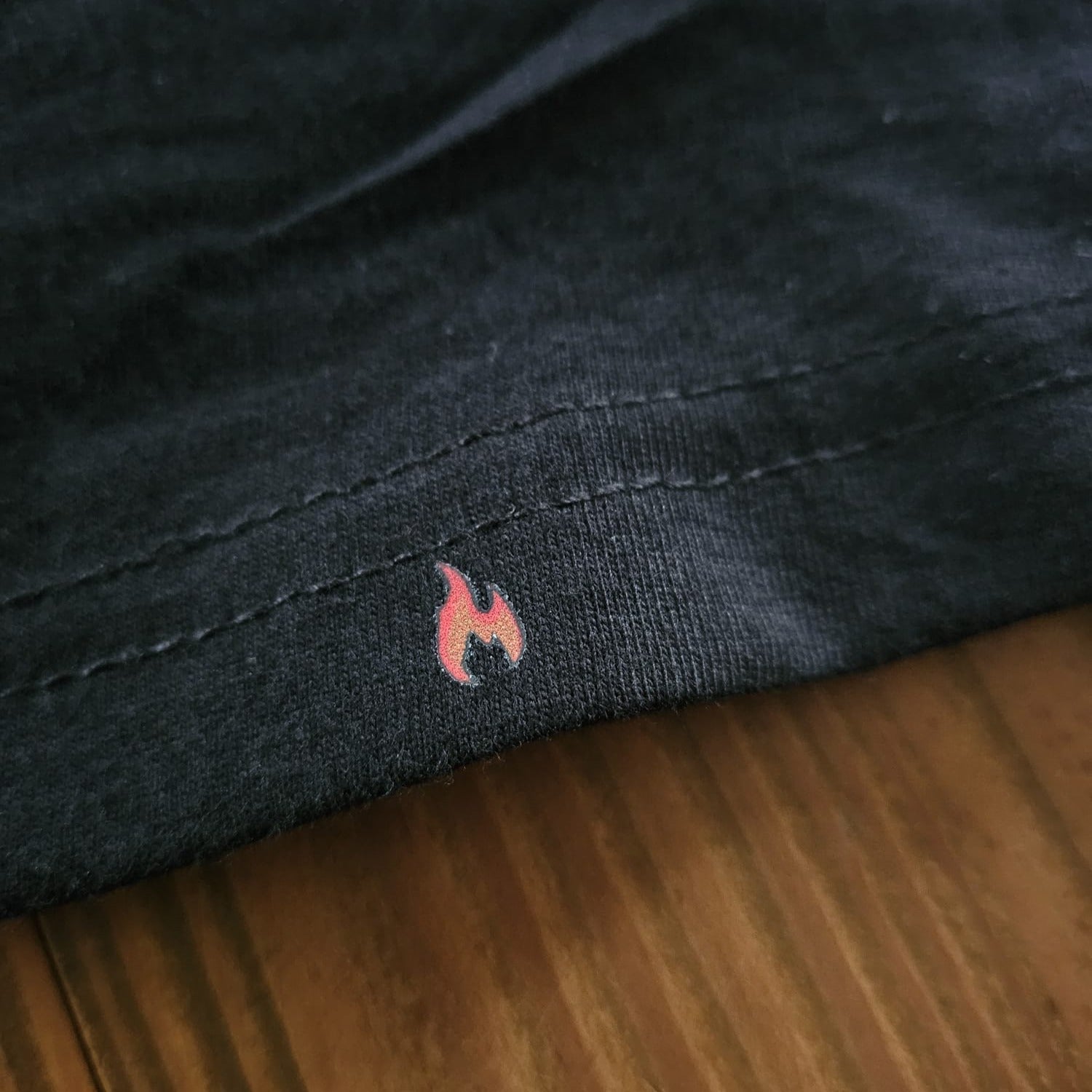Nahaufnahme des unteren T-Shirt-Saums mit einer kleinen, dezent eingedruckten roten Flamme auf einer Holzoberfläche.