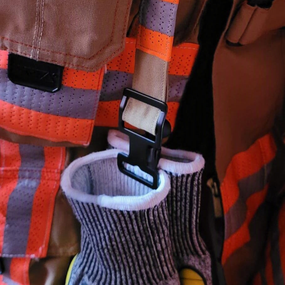 "Schutzhandschuhe eingehängt in schwarzen Taktik-Clip des Feuerwehr-Handschuhhalters an Einsatzjacke mit orangenen Reflexstreifen – griffbereit für die Technische Hilfeleistung"