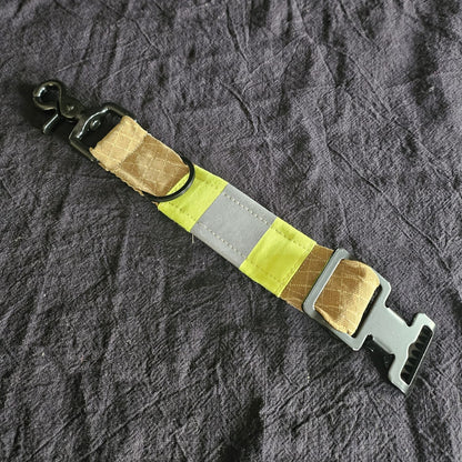 "Draufsicht auf Feuerwehr-Handschuhhalter aus sandfarbenem Aramid-Einsatzstoff mit neongelb-silbernem Reflexstreifen, schwarzem Scherenkarabiner und Metallschnalle – 25 x 3 cm Upcycling-Unikat