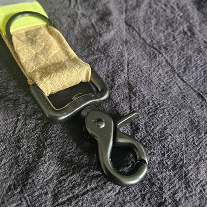 "Schwarzer Scherenkarabiner und Schlüsselring am sandfarbenen Feuerwehr-Handschuhhalter aus Aramid-Einsatzstoff mit neongelben Reflexstreifen – Detailansicht der Befestigungsmechanik"

