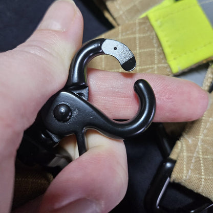 "Hand öffnet schwarzen Scherenkarabiner aus Zinklegierung – Nahaufnahme der Federmechanik des Feuerwehr-Handschuhhalters vor sandfarbenem Aramid-Einsatzstoff mit neongelben Reflexstreifen"
