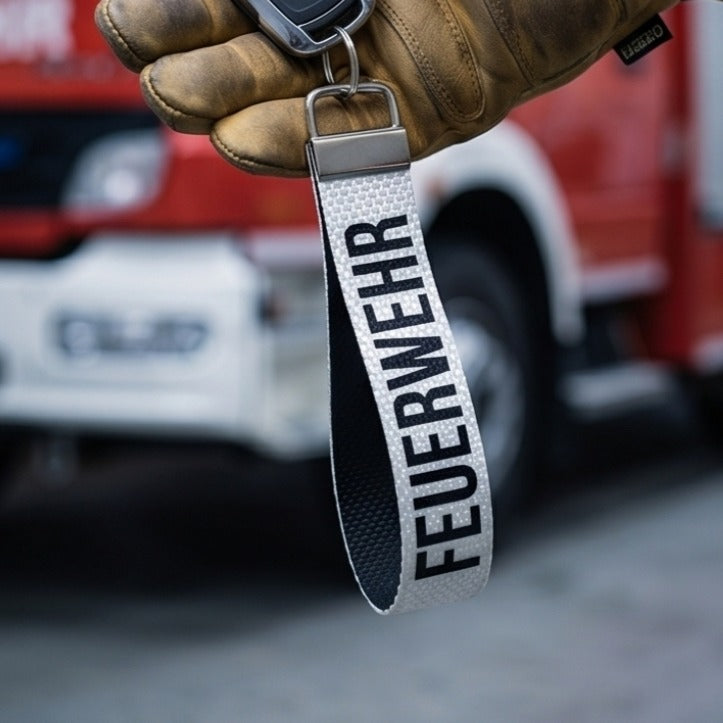 "Weißer Feuerwehr-Schlüsselanhänger aus Feuerwehrschlauch mit schwarzer FEUERWEHR-Aufschrift vor rotem Feuerwehreinsatzfahrzeug – Lifestyle-Aufnahme"
