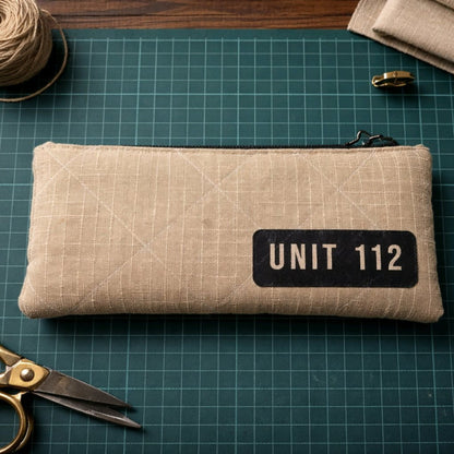 Flach liegendes Etui UNIT 112 aus beigem Upcycling-Feuerwehrschlauch mit schwarzem Logo-Aufdruck auf einer grünen Schneidematte.